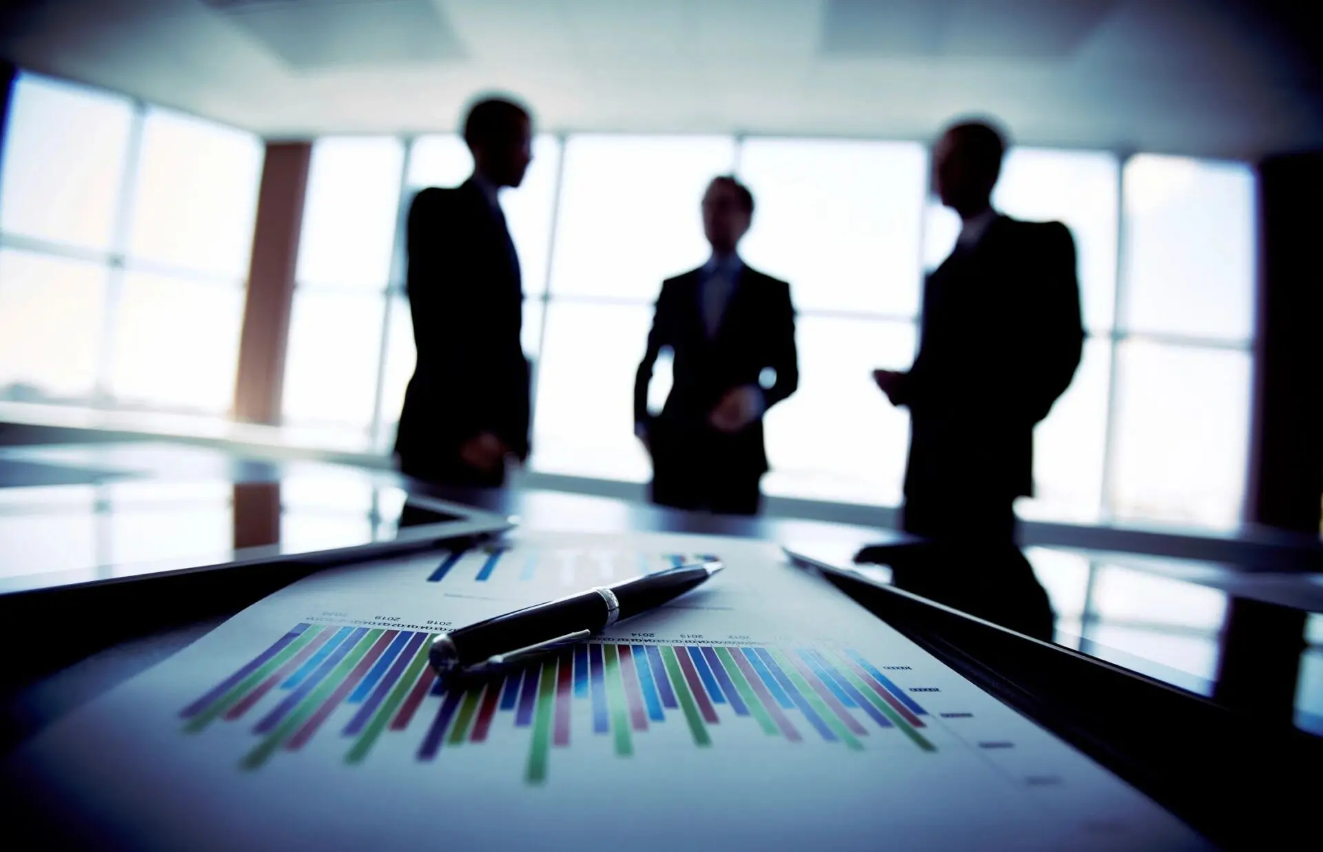 Laptop and Financial Document With Pen at Workplace on Background of Three Business Partners Interacting